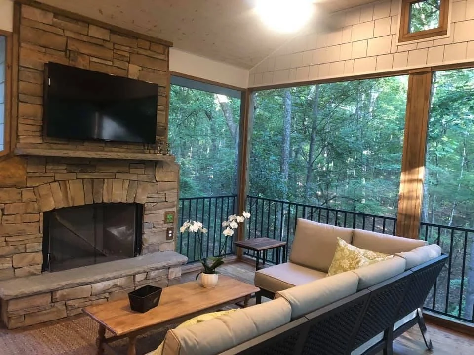 Screened porch with stacked stone fireplace, TV mount, large windows, and black metal railings overlooking wooded landscape